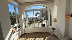 A room with a large arched window, wooden flooring, a small round table with a plant, and a white wall with a decorative item.