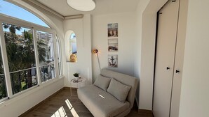 A room with a sofa, a small table with a plant, artwork on the wall, and a large window with a view of palm trees.