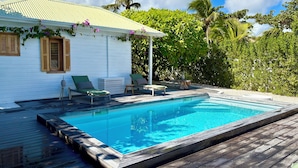A poolside area with a wooden deck, a white building with a green roof, and lush greenery in the background. - VILLA EDENLY ORIENT BAY (saint martin)