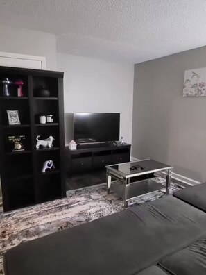 Uma sala de estar moderna com um sofá escuro, uma mesa de centro de vidro, um rack preto com uma televisão de tela plana e uma estante decorativa com diversos objetos.