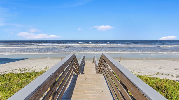 Ein hölzerner Steg führt zu einem Strand, im Hintergrund sind Wellen zu sehen.