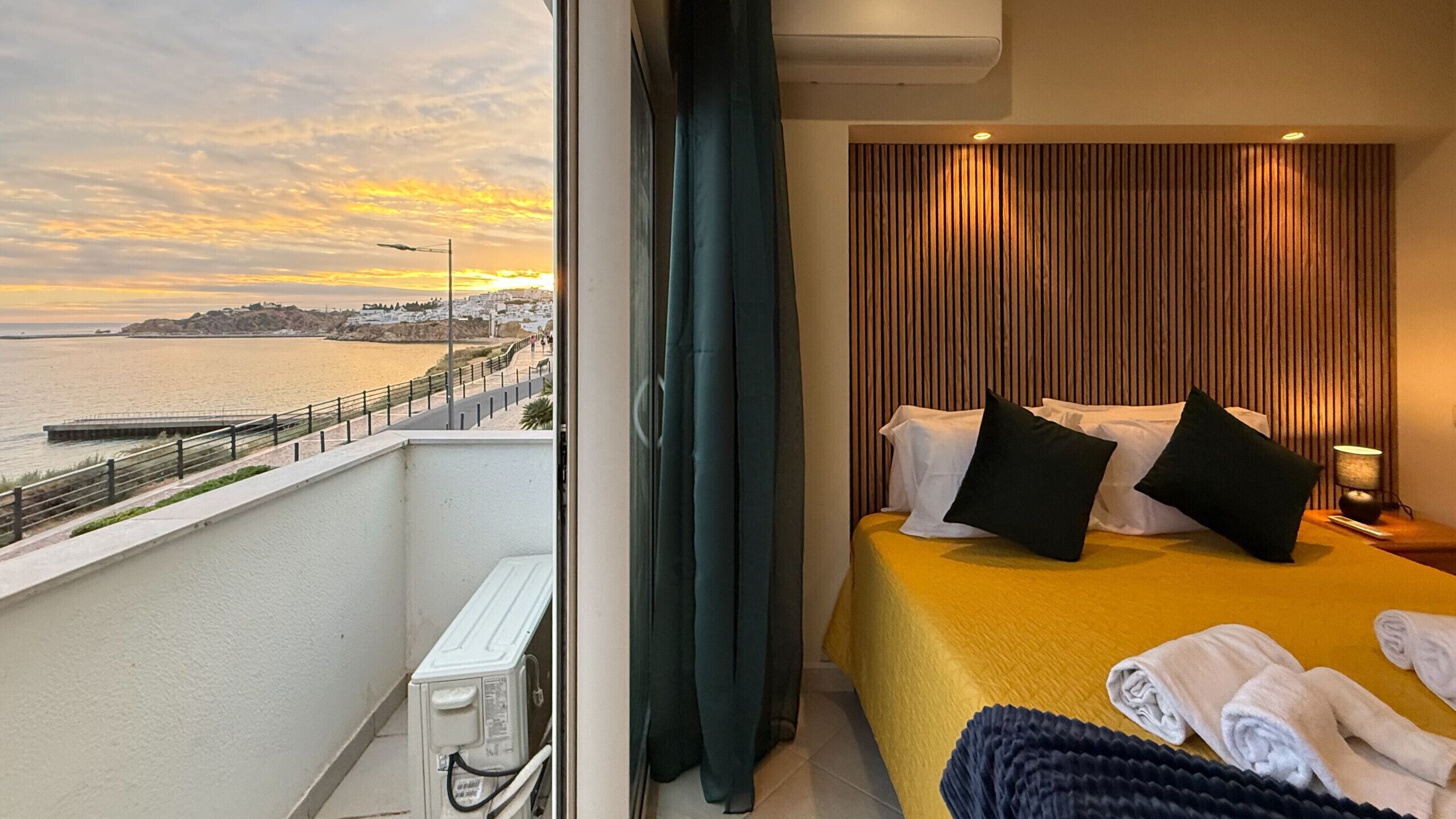 A hotel room with a bed, pillows, a nightstand, a lamp, and a view of the sea and a coastal town.