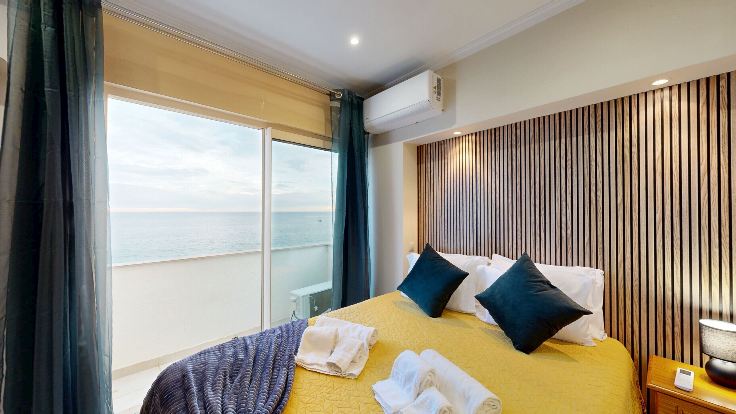 A hotel room with a bed, a nightstand, a lamp, a window with curtains, and an air conditioning unit.