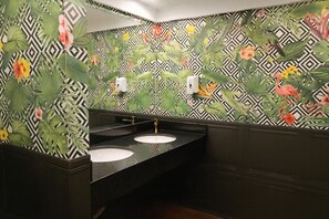 Un baño con dos lavamanos, un espejo y un secador de manos. Las paredes están decoradas con un estampado floral tropical.