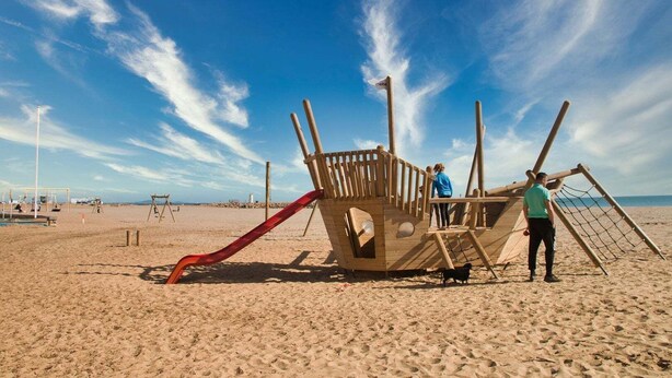 Eine Holzspielkonstruktion an einem Sandstrand mit einer roten Rutsche und einem Bereich mit Netz.