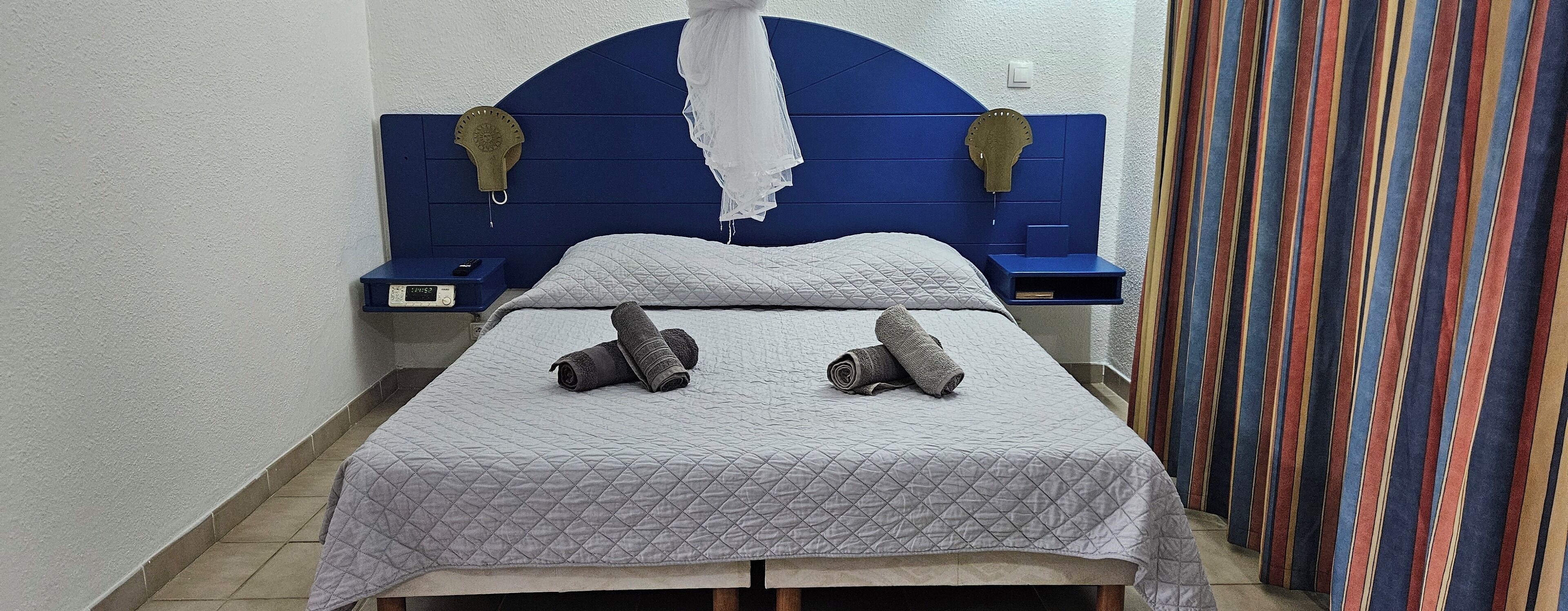 A single bed with a quilted grey bedspread, two rolled-up towels, a blue headboard with decorative elements, a small bedside table with a digital clock, and a striped curtain.