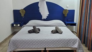 A single bed with a quilted grey bedspread, two rolled-up towels, a blue headboard with decorative elements, a small bedside table with a digital clock, and a striped curtain.