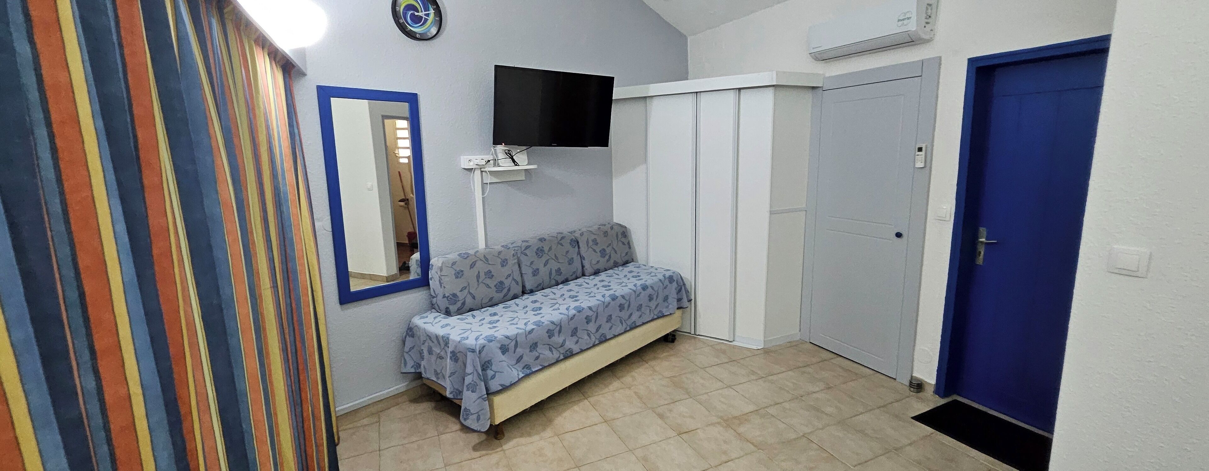 Un petit couloir étroit avec un canapé, un miroir, un téléviseur et une porte bleue.