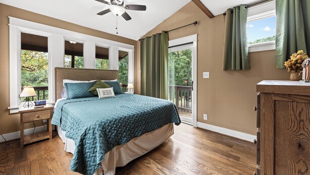 A bedroom with a bed, a nightstand, a ceiling fan, a window with curtains, and a view of trees.