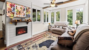 A cozy living room with a fireplace, large windows, a ceiling fan, and a comfortable seating area.