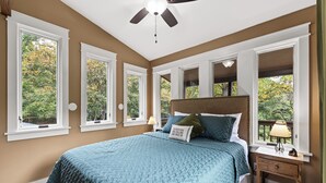 A bedroom with a large bed, multiple windows, a ceiling fan, and a nightstand with a lamp.