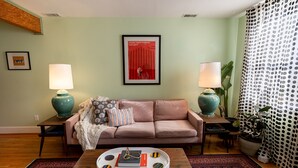 A living room with a sofa, two side tables with lamps, a coffee table with decorative items, a framed picture on the wall, and a patterned curtain. - Wyatt s Saloon Guest House 2 (Baltimore)