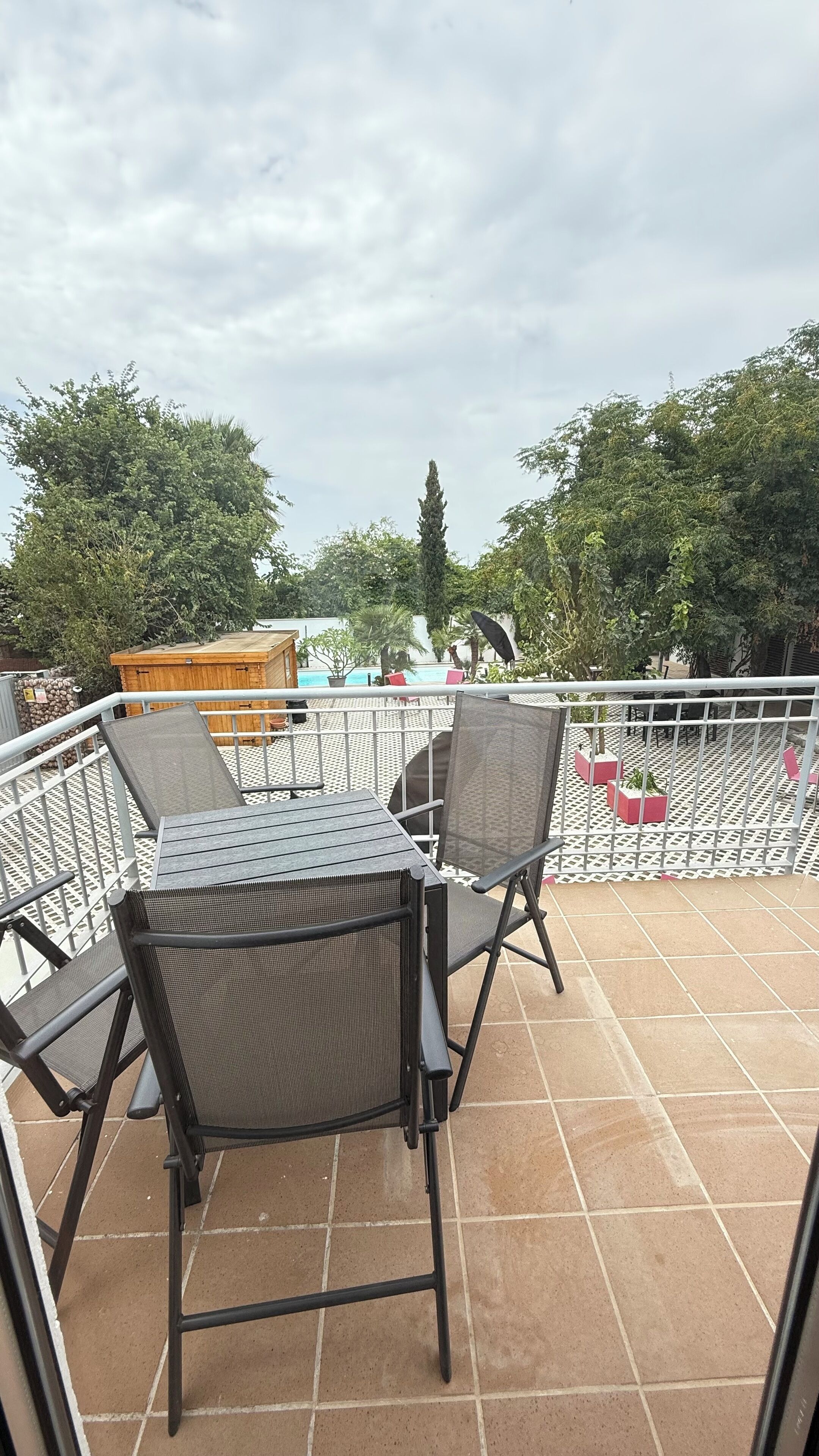 Une terrasse avec une table et des chaises, une structure en bois, et une vue sur des arbres et un bâtiment en arrière-plan.