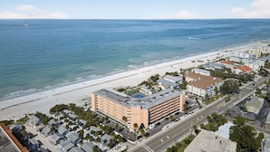 Ein Luftbild eines Küstenhotels mit Strandlage, das ein großes Gebäude mit Pool auf dem Dach, angrenzendem Parkplatz und Blick auf den Ozean zeigt.