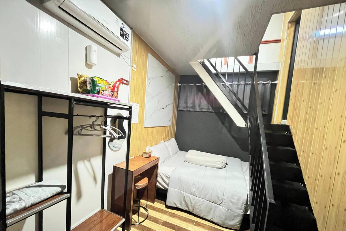 A compact room with a bunk bed, a small wooden table, a chair, and a desk. There is a staircase leading to an upper level.