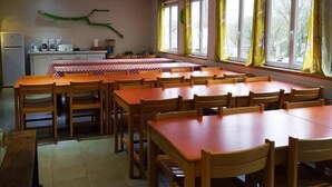 A dining area with wooden tables and chairs, a kitchen area with appliances, and large windows with curtains. - Gite D'étape port de Carhaix (Carhaix-Plouguer)