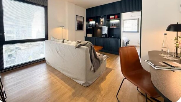 A modern living room with a sofa, a coffee table, and a desk with a chair. There is a large window with blinds, a bookshelf, and a wall-mounted shelf with decorative items.