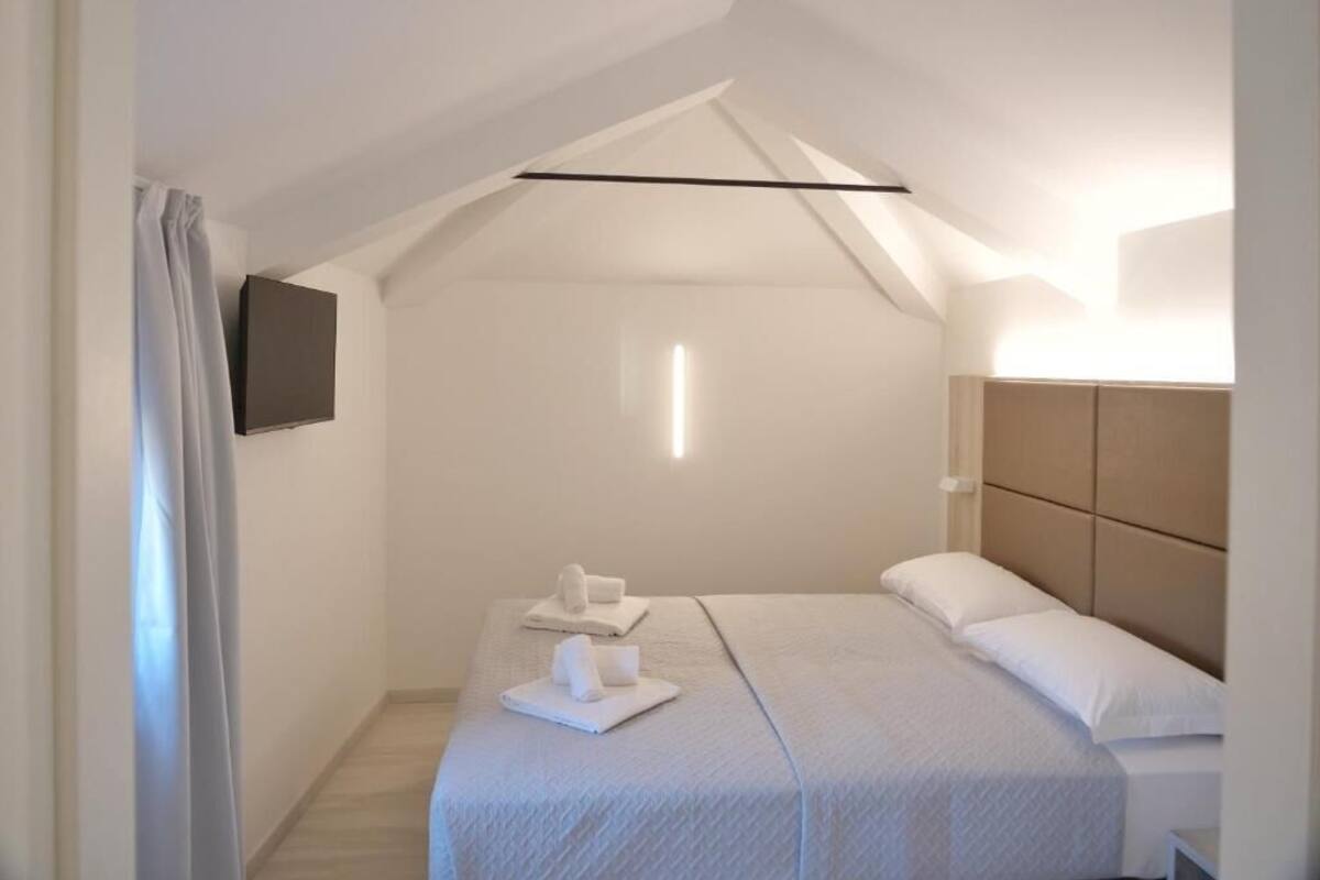 A modern bedroom with a sloped ceiling, a flat-screen TV mounted on the wall, and a neatly made bed with towels.
