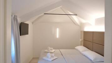 A modern bedroom with a sloped ceiling, a flat-screen TV mounted on the wall, and a neatly made bed with towels.