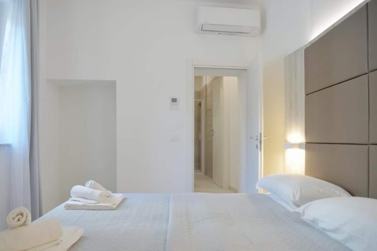 A hotel room with a bed, a bedside table, a wall-mounted air conditioner, and a door leading to another room.