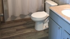 Un baño con piso de madera, inodoro blanco, cortina de ducha y lavamanos con encimera.
