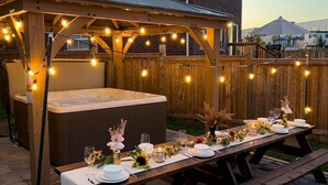 Et udendørs spiseområde med jacuzzi, pergola i træ, lyskæder og et spisebord dækket til et måltid.