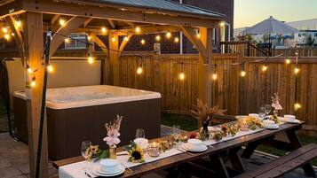 En utomhusplats för måltider med en bubbelpool, en träpergola, ljusslingor och ett picknickbord som är dukat för en måltid.