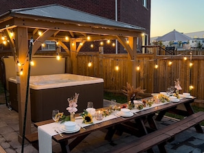 En utomhusplats för måltider med en bubbelpool, en träpergola, ljusslingor och ett picknickbord som är dukat för en måltid.