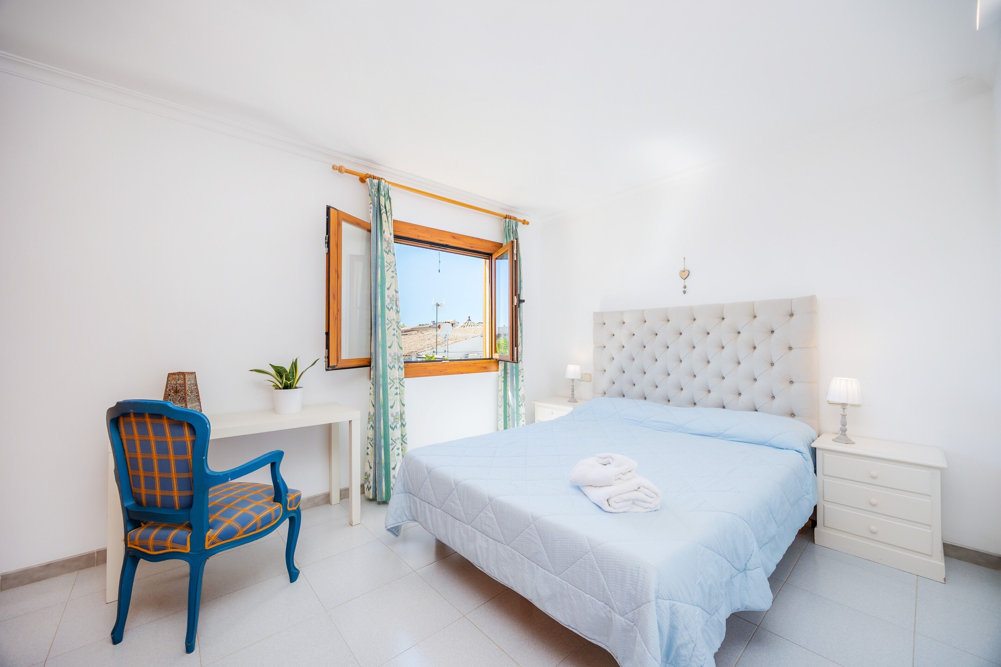 A bedroom with a bed, a chair, a desk, a nightstand, a window with curtains, and a view of buildings outside.