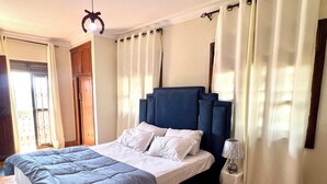 A bedroom with a large bed, a bedside table with a lamp, and curtains on the windows. - Beautiful 2-bedroom apartment in Muyenga city center in Kampala with Lake View (Kampala)