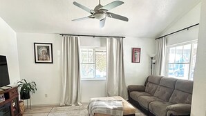 Ein Wohnzimmer mit Deckenventilator, einer Couch, einem Couchtisch, einem Fernseher und einem Fenster mit Vorhängen.