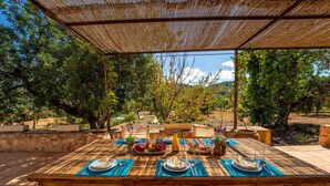 An outdoor dining area with a wooden table set for four, surrounded by chairs, under a woven shade structure.