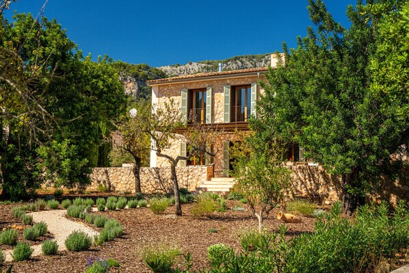 A Mediterranean-style villa with a stone facade, a wooden balcony, and a garden with various shrubs and a gravel path.
