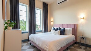 Une chambre à coucher comprenant un lit, une commode, une plante en pot, deux fenêtres avec des rideaux et un climatiseur fixé au mur.