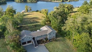 Une grande maison avec un garage, entourée d’une pelouse bien entretenue et d’arbres, située près d’un lac.