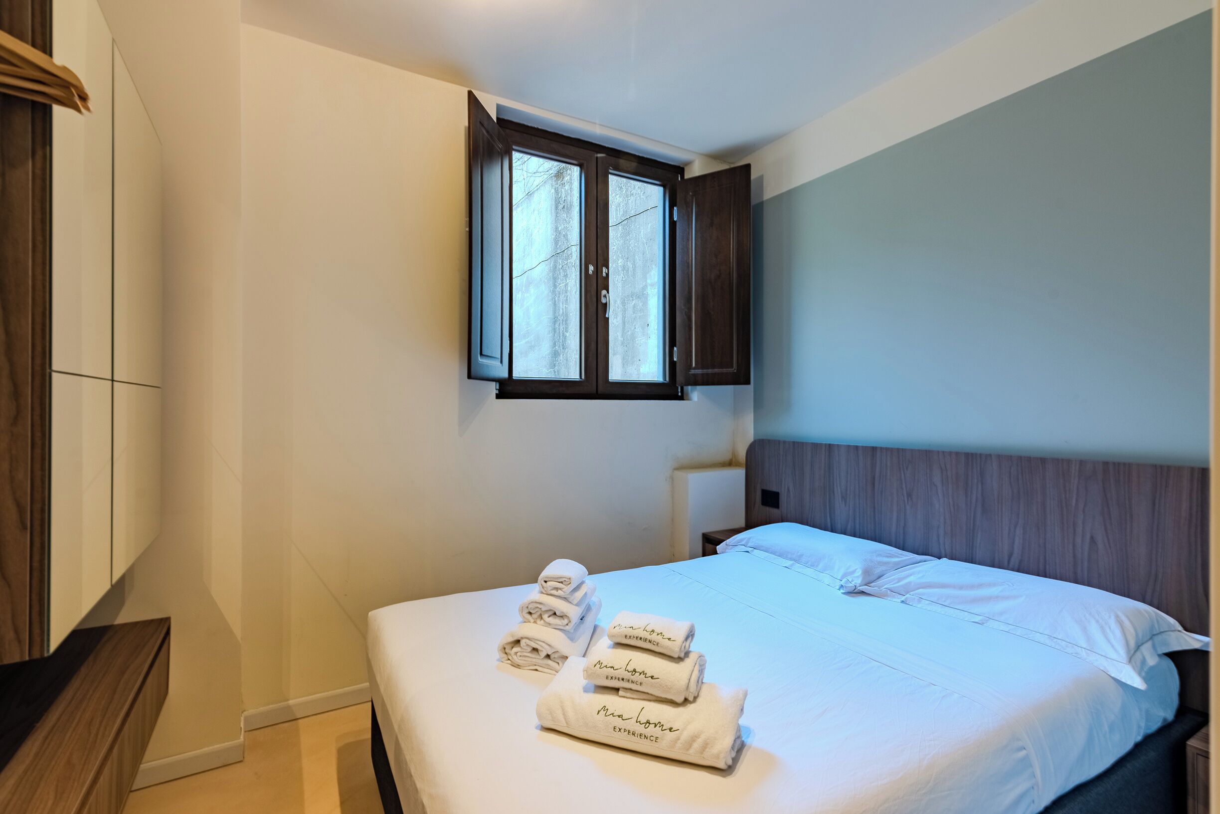 A neatly made bed with white linens and towels, a wooden headboard, and a window with dark frames.
