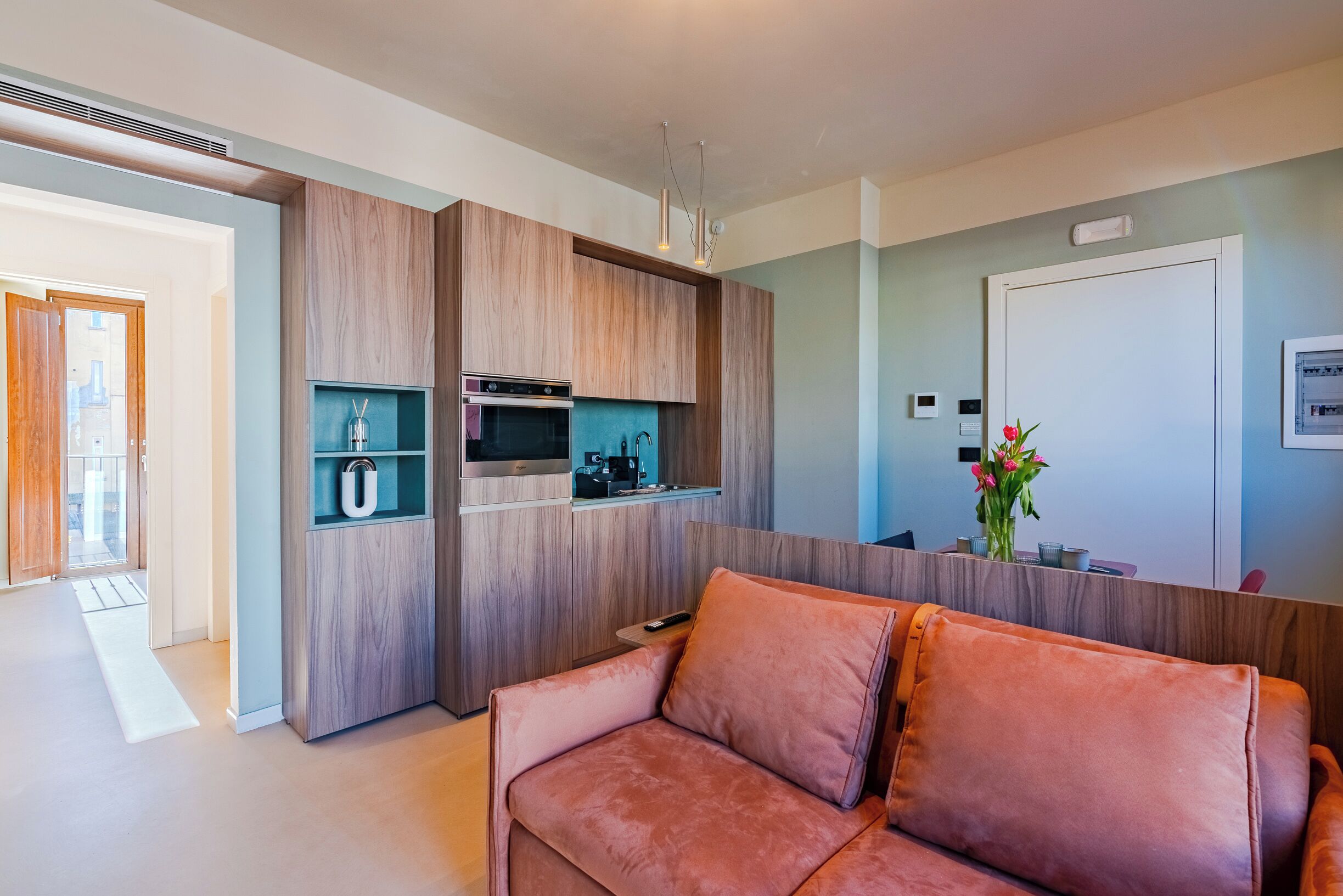 A modern living room with a sofa, wooden cabinetry, and a kitchen area with a microwave and a coffee machine.