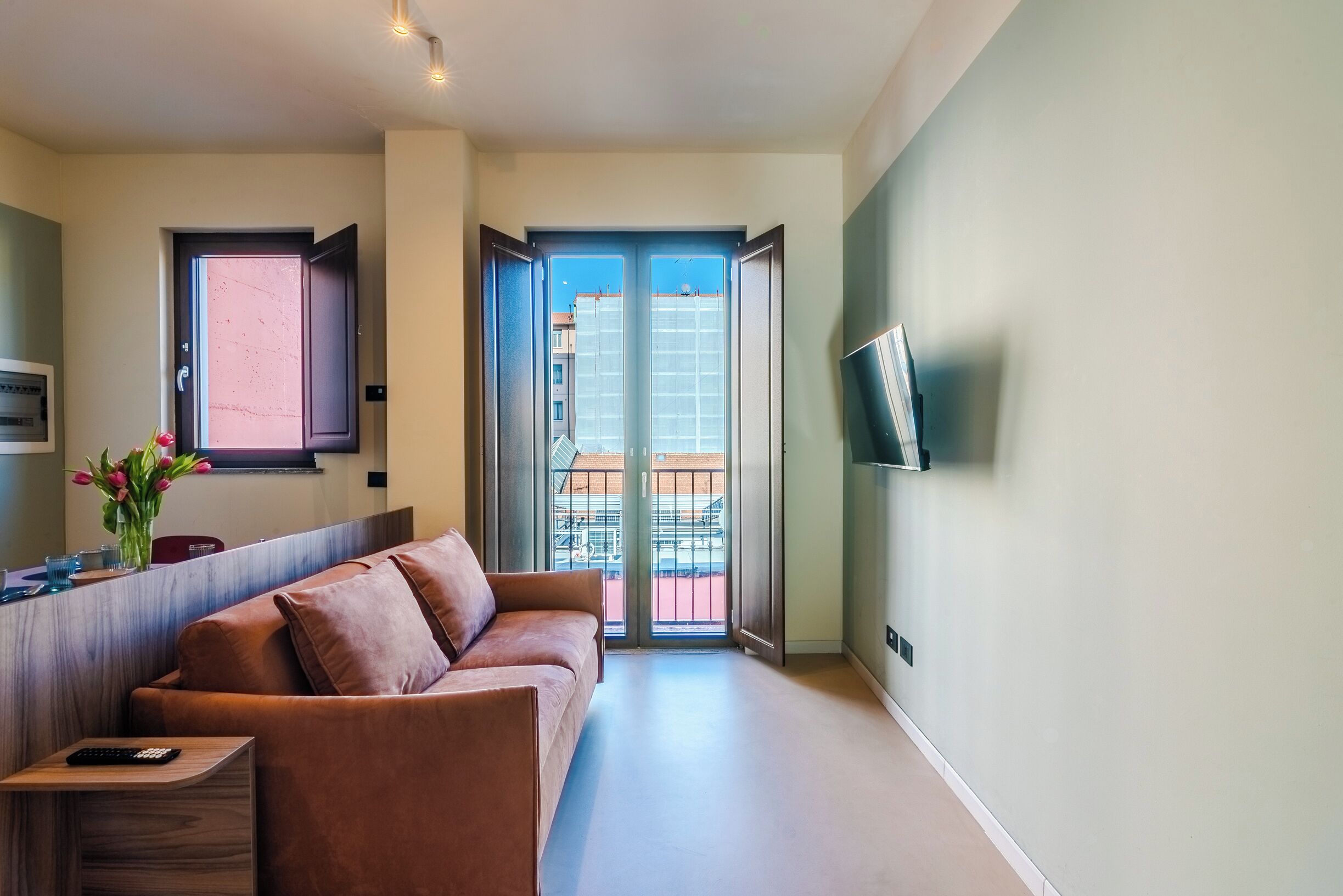 A modern living room with a sofa, a small table with a remote control, a TV mounted on the wall, and a large sliding glass door leading to an outdoor balcony.