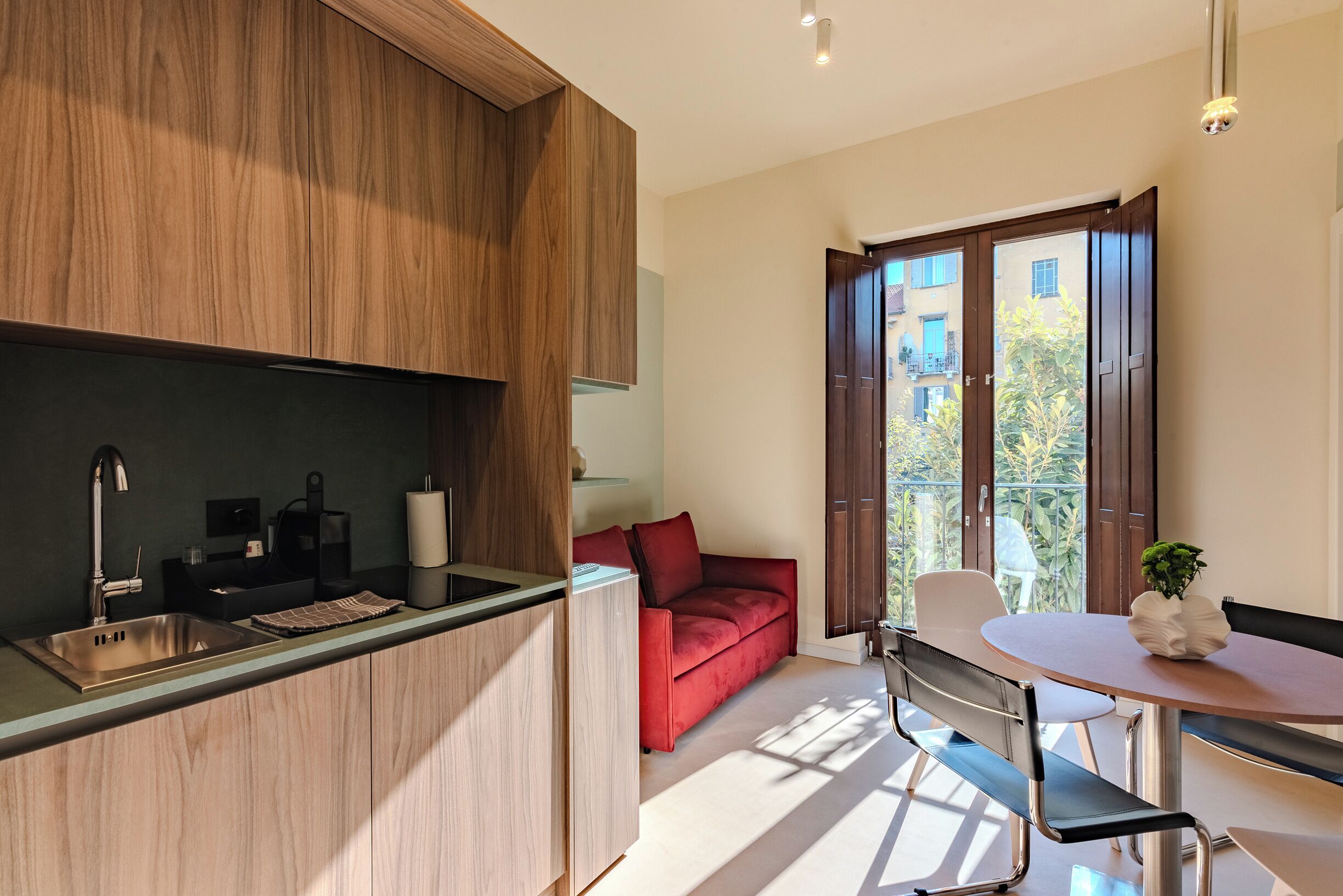 A modern kitchen with a built-in sink and wooden cabinets, a dining area with a table and chairs, and a red sofa in a bright room with large windows.