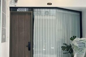 Een houten deur met een zwarte klink, een witte muur met een nummerplaatje, en een grote potplant aan de rechterkant.