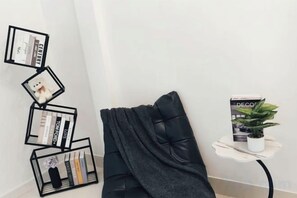 Un coin de pièce avec un fauteuil noir recouvert de tissu, une petite table d’appoint blanche sur laquelle est posée une plante en pot, et un mur orné d’une collection d’objets encadrés.