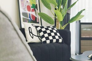 En svart soffa med en rutig kudde och en enfärgad kudde, en stor grön växt och en inramad abstrakt målning i ett rum med ett fönster.