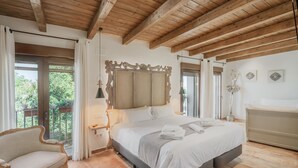Une chambre avec un grand lit, des poutres apparentes en bois au plafond, une chaise et un balcon agrémenté de rideaux.