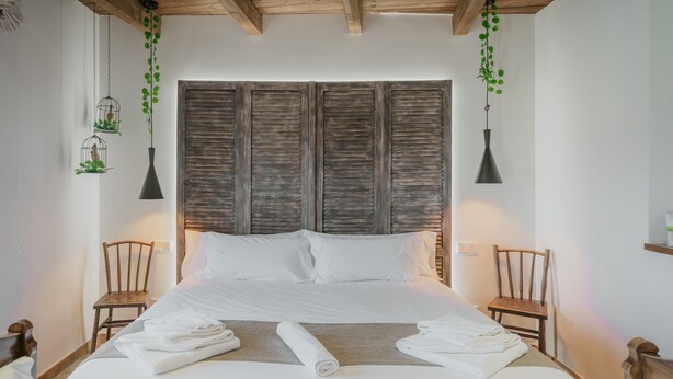 Un lit bien fait, avec du linge de lit blanc, deux chaises en bois et une tête de lit ornée d’un motif texturé.