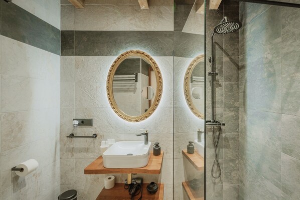 Un baño con lavamanos blanco, tocador de madera, espejo con marco dorado, toallero y ducha.