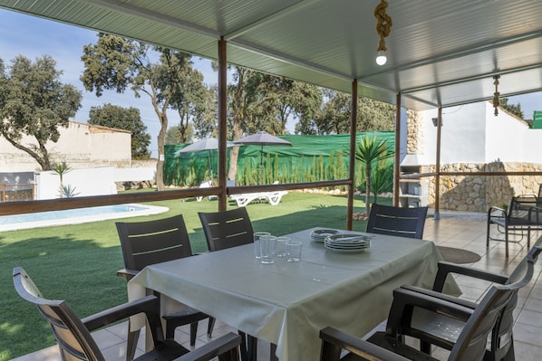 An outdoor patio area with a table set for dining, surrounded by chairs, and a view of a swimming pool and greenery.