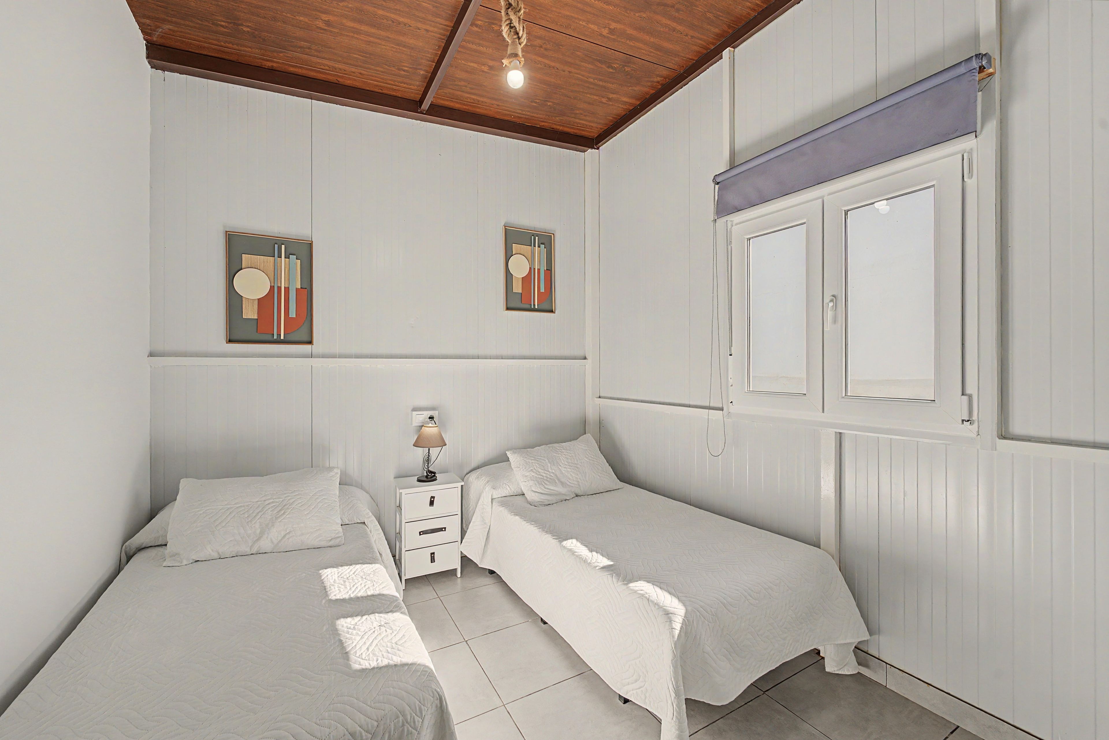 A bedroom with two single beds, a nightstand with a lamp, and a window with blinds.