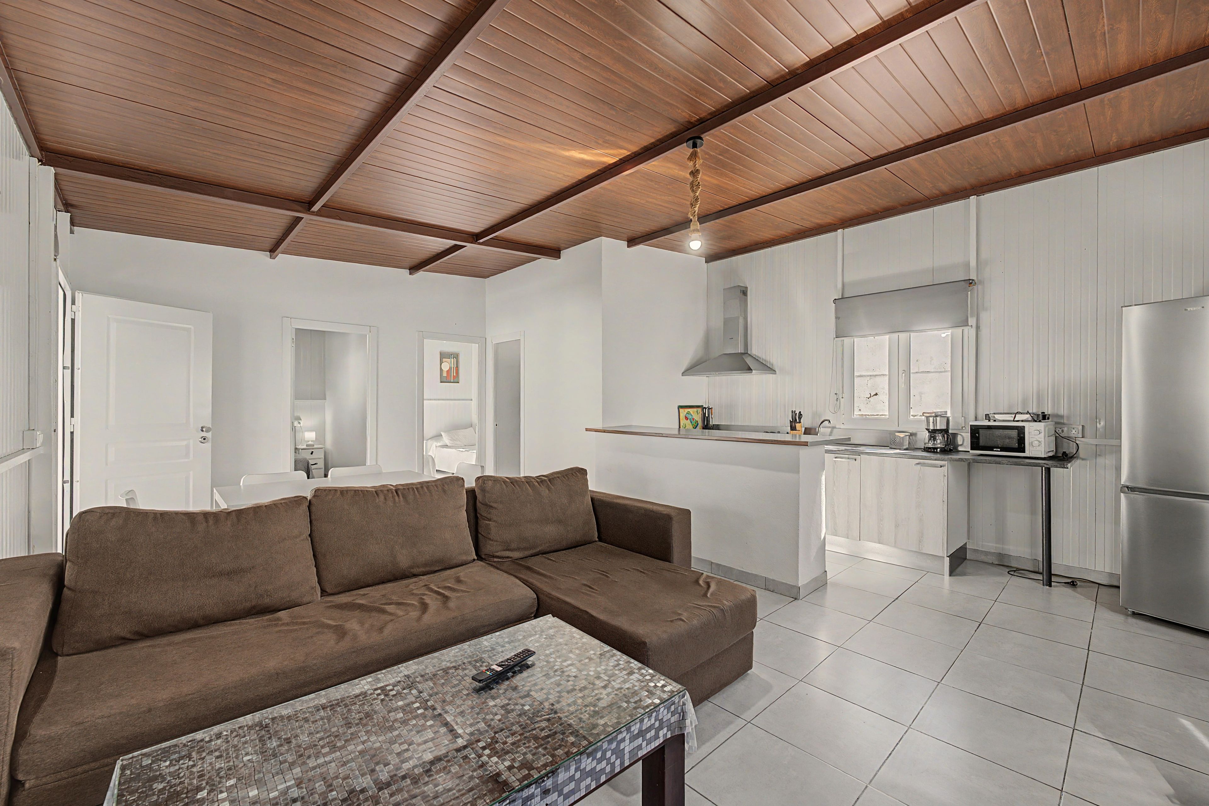 Una sala de estar moderna con un sofá marrón, una mesa de centro de vidrio y una cocina con armarios blancos y electrodomésticos de acero inoxidable.
