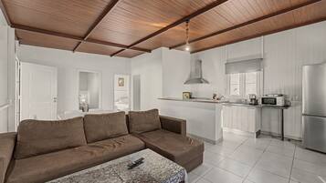 Una sala de estar moderna con un sofá marrón, una mesa de centro de vidrio y una cocina con armarios blancos y electrodomésticos de acero inoxidable.