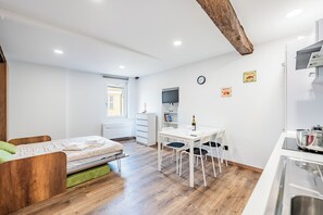 Une pièce de taille modeste comprenant un lit, une petite table à manger avec des chaises, un téléviseur fixé au mur et un coin cuisine.